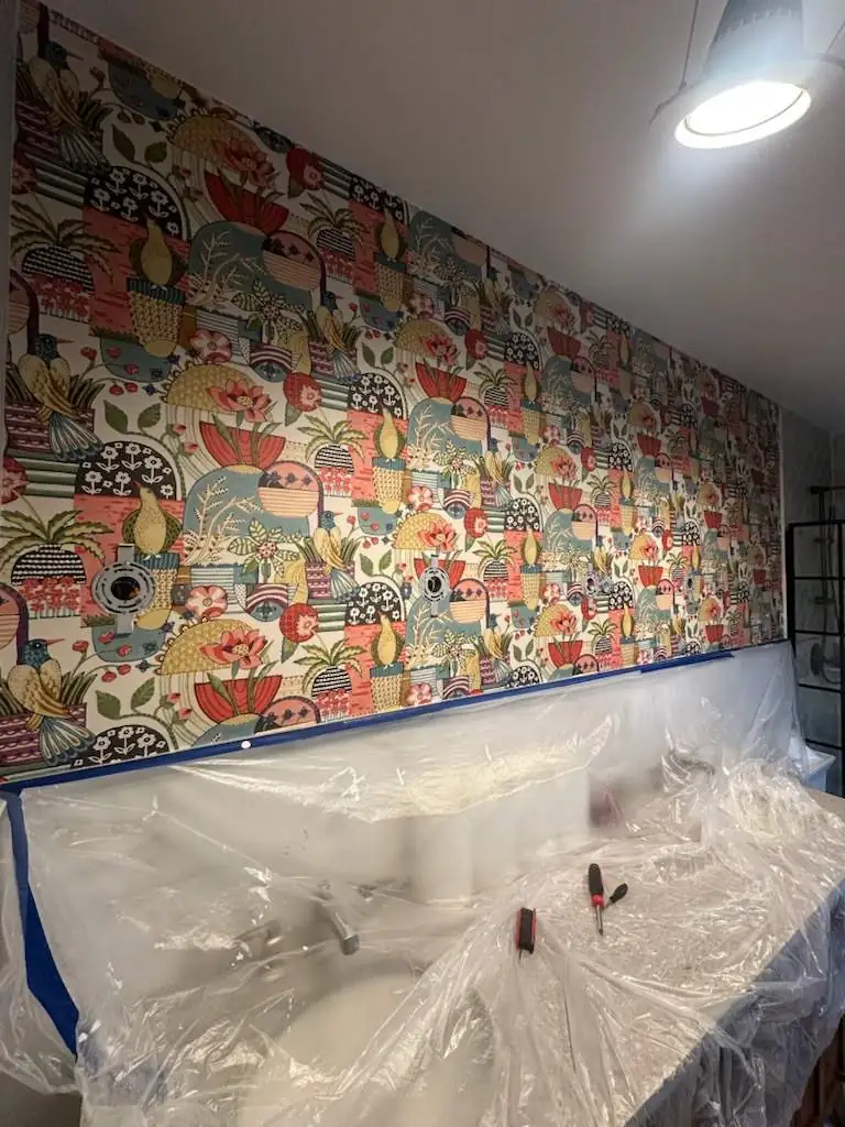 A wallpaper install in progress: paper hung, sconces removed and electrical boxes cut out, vanity covered in plastic
